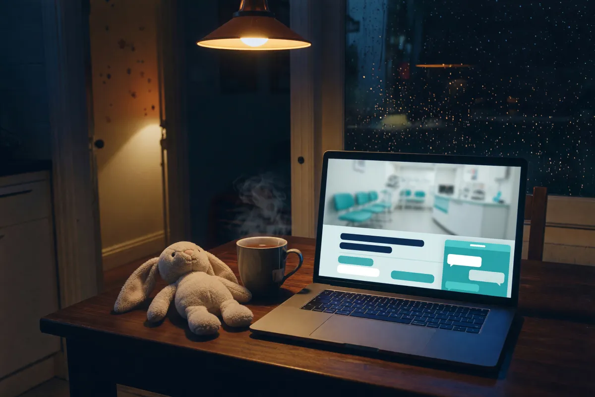 Laptop on a kitchen table at night showing a clinic website with a chat panel open, a child's door ajar behind